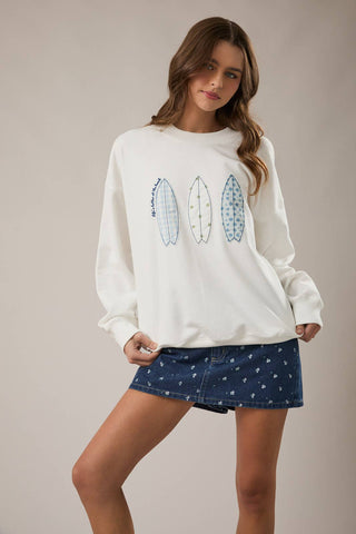 Woman wearing coastal surfboard graphic sweatshirt with embroidered surfboard design.